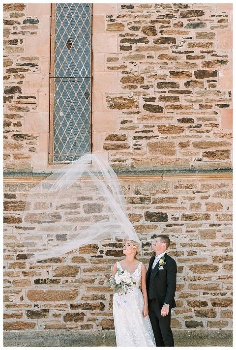 bride and groom watching veil fly in wind outside church